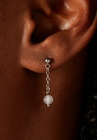 Les boucles d'oreilles en chaîne argentée présentent un petit design en forme de balle avec une perle blanche translucide, suspendue à un clou dans une vue rapprochée de l'oreille.