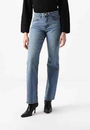 Kõrge vöökohaga sinised denim püksid sirge lõikega, heledas toneeringus ja klassikalise viie tasku disainiga; kombineeritud mustade kõrgete kontsadega kingadega.