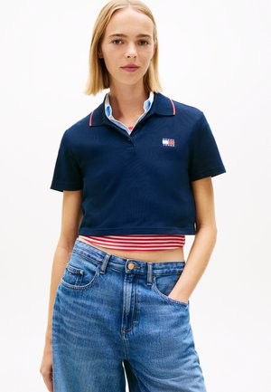 Noor naine kannab tumesinist lühikest polo särki punase-valge triibulise topi ja kõrge vöökohaga siniste teksapükstega, üks käsi taskus.