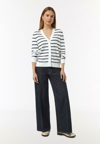Weißer Cardigan mit marineblauen Streifen, V-Ausschnitt und Knopfleiste. Kombiniert mit weit geschnittenen, dunklen Jeans aus Denim, die orangefarbene Nähte und schwarze Schuhe aufweisen.