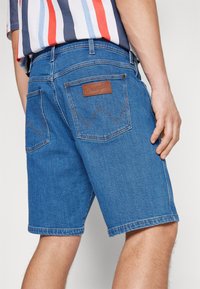 Pantalones cortos de mezclilla Wrangler en azul medio, con un parche de cuero marrón en la parte trasera, costuras distintivas y dobladillo vuelto.