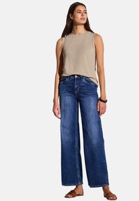 Top beige senza maniche con dettagli a borchie, abbinato a jeans in denim blu a gamba larga. I jeans presentano un lavaggio chiaro e una vestibilità rilassata.