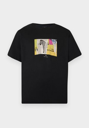 Ung mand med krøllet sort hår iført en sort t-shirt med grafik, løse blå jeans og beige loafers, stående mod en ensfarvet hvid baggrund.