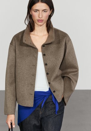 Frau trägt eine braune Wolljacke über einem weißen Top mit einem blauen Pullover um die Taille gebunden und dunkle Jeans, steht mit einer Hand in der Tasche.