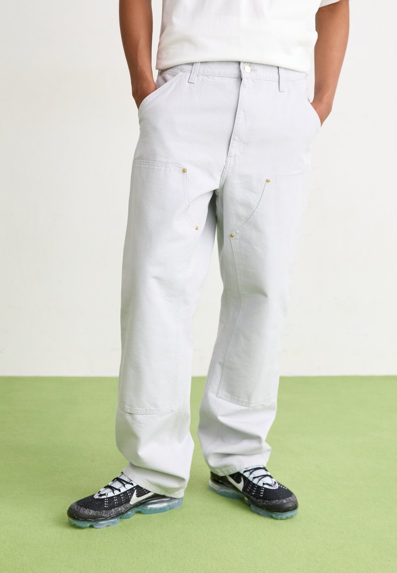 Light gray pants with a relaxed fit, featuring front pockets and gold-tone accents. Paired with black sports shoes and a white t-shirt.