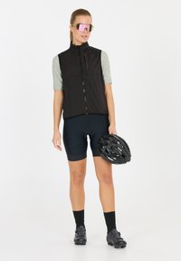 Schwarzige Radweste über einem hellgrünen T-Shirt mit kurzen Ärmeln, kombiniert mit navyblauen Shorts. Das Model hält einen schwarzen Helm und trägt pinke Sonnenbrillen.