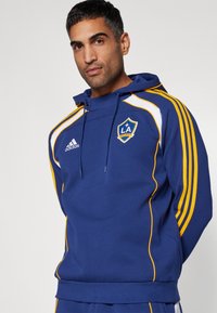 Blue hoodie made of cotton blend featuring a front pocket, yellow accents, Adidas logo, LA Galaxy emblem, and a drawstring hood.