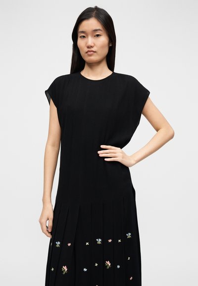 Robe noire sans manches avec un col rond, dotée de plis verticaux et de motifs floraux brodés en couleurs pastel le long de l'ourlet.