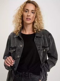 Czarna dżinsowa kurtka z zapinanymi kieszeniami, noszona na czarnym swetrze. Model trzyma okulary przeciwsłoneczne, ma falowane włosy do ramion i neutralne tło.