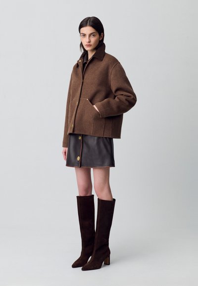 Veste en laine marron avec un col et des boutons sur le devant, assortie à une mini-jupe en cuir noir et de longues bottes en suède marron à talons carrés.