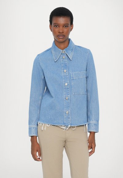 Light blue denim jacket with a shirt collar, front button closure, a patch pocket, and frayed hem. Styled with beige pants.