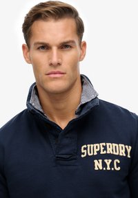 Polo de color azul marino con cuello a cuadros, con tapeta de botones y logo "SUPERDRY N.Y.C" en color crema, hecho de material de algodón.