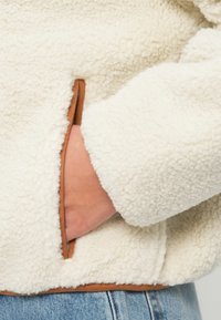 Bojna fleec jakna boje kreme s teksturiranom površinom, koja ima smeđu kožnu akcentu oko džepa i struka.