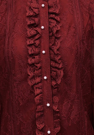 Βαθύ κόκκινο δαντελένιο ύφασμα με κάθετο βολάν διακοσμητικό και μια σειρά από λευκά μαργαριταρένια κουμπιά στο κέντρο.