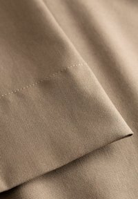 Lähikuva beigestä kankaasta, jossa näkyy vinokudoksen kuvio ja siisti saumalinja yläosassa.