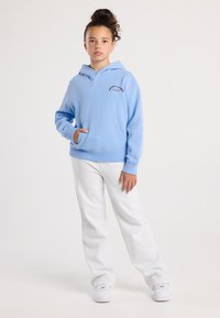 Giovane con una mano in tasca, che indossa una felpa leggera blu, pantaloni bianchi e sneakers bianche su uno sfondo neutro.