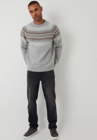 Grijze gebreide trui met een geometrisch patroon in wit, bruin en lichtgrijs over de borst, gecombineerd met donkere spijkerbroeken en grijze sneakers.