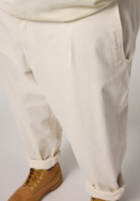 Pantaloni larghi di un beige chiaro, con tasche laterali e realizzati in un tessuto testurizzato. Orlati alla caviglia, con stivali marroni stringati visibili.