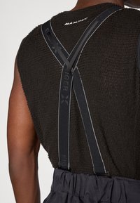 Czarna, teksturowana, bezrękawnikowa odzież z regulowanymi czarnymi szelkami z refleksyjnymi akcentami i logo. Ciemne spodnie widoczne poniżej.