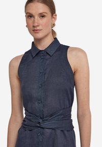 Mörkblå ärmlös klänning med knapprad fram, krage och en knuten midjedetalj. Tillverkad av mjukt, texturerat tyg.