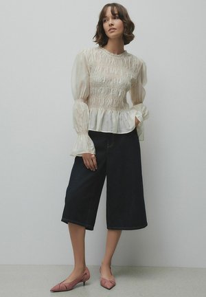 Sieviete ar īsiem brūniem matiem, tērpusies krēmkrāsas blūzē ar rukām, platām tumšām ceļgalu garuma biksēm un rozā zemu papēžu kurpēm, stāvot pret vienkāršu sienu.