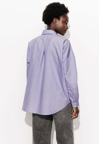 Chemise à manches longues rayée violet et blanche avec une texture lisse, dotée d'un col pointu, de poignets à boutons et d'un ourlet arrondi.