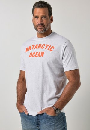 Mann mit Bart, trägt ein graues "Antarctic Ocean"-T-Shirt, blaue Jeans und eine schwarze Uhr, steht mit den Händen in den Taschen vor schlichtem Hintergrund.