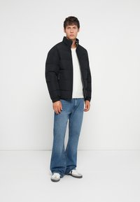 Giovane uomo che indossa un giubbotto a vento nero, un maglione bianco, jeans a gamba larga blu e sneaker bianche, in piedi davanti a uno sfondo bianco semplice.