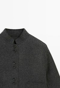 Cappotto di lana grigio scuro con colletto alto, sei bottoni neri sul davanti e una tasca sul petto.