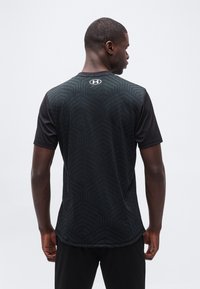 Άνδρας γυρισμένος προς τα πίσω, φοράει μαύρο αθλητικό μπλουζάκι με κοντά μανίκια, διακοσμημένο με διακριτικά γεωμετρικά σχέδια και λευκό λογότυπο Under Armour στο λαιμό.