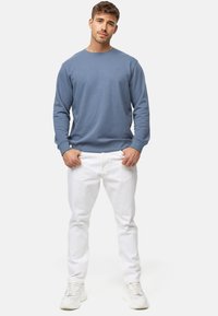 Sudadera de cuello redondo azul en tejido suave, combinada con jeans blancos. Detalle del logo visible en el pecho, el modelo se encuentra en un fondo neutro.