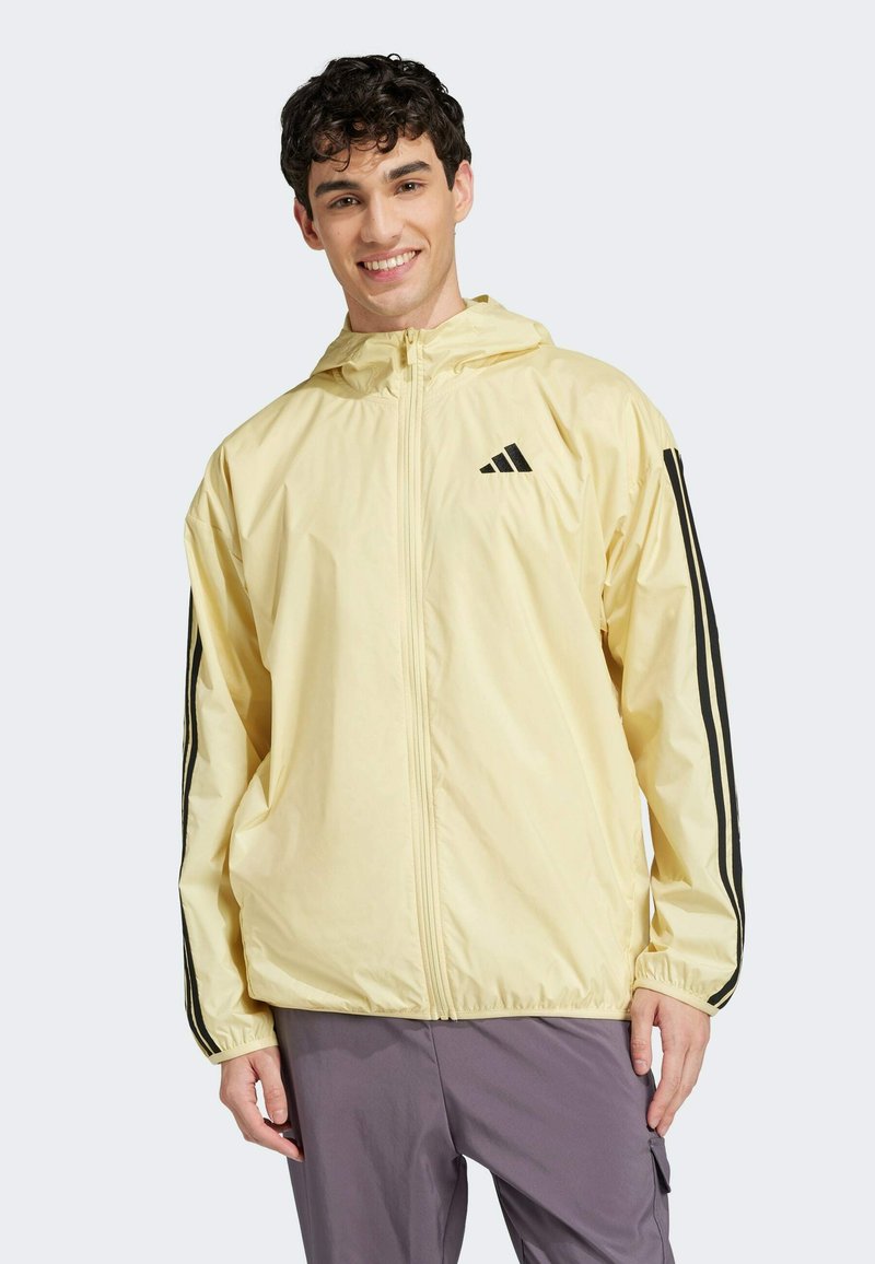 Lys gul vindjakke med frontlynlås, sort Adidas-logo og sorte sidelister. Har hætte og elastiske manchetter.