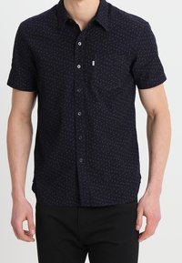 Kortärmad, mörkblå skjorta med knappar och ett subtilt geometriskt mönster, en bröstficka och en slät tygtextur.