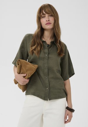 Femme aux longs cheveux ondulés portant une chemise kaki à manches courtes boutonnée, un pantalon blanc, tenant une veste marron pliée et portant un bracelet noir.