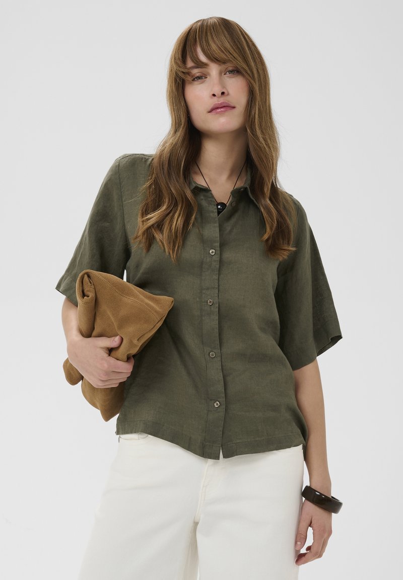 Femme aux longs cheveux ondulés portant une chemise kaki à manches courtes boutonnée, un pantalon blanc, tenant une veste marron pliée et portant un bracelet noir.