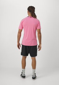 Mies, który ma dredy, stoi tyłem, ubrany w różową koszulkę sportową, czarne szorty sportowe, białe skarpetki Adidas i czarne buty Adidas.