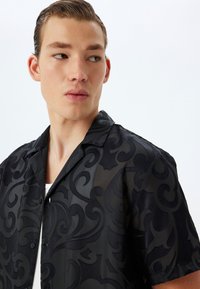 Camisa negra de manga corta con un sutil patrón en relieve de espirales, fabricada en un tejido ligero y con collar clásico. Cierre de botón en la parte delantera.