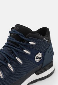 Zapato de senderismo de tela azul con acentos reforzados en negro, ojales plateados y un logo de árbol. Presenta un tejido texturizado y una suela de goma resistente.