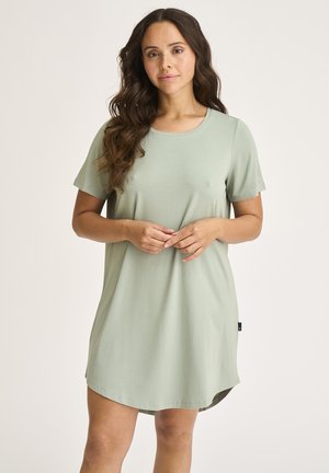 Kvinde med langt, bølget hår iført en løs, salviegrøn kjole med korte ærmer, stående foran en ensfarvet lys baggrund.