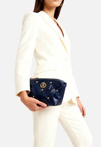 Borsa trapuntata in ecopelle blu navy con borchie dorate e logo circolare, con chiusura a zip e forma compatta, tenuta contro un abito bianco.