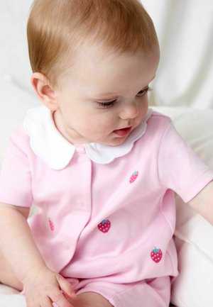 Bebeluș cu păr șaten deschis care poartă un costum roz cu guleraș alb și mici imprimeuri cu căpșuni, uitându-se în jos și întinzând mâna înainte.