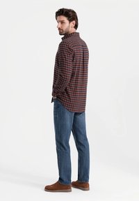 Camicia a quadri marrone e navy, maniche lunghe, colletto button-down, abbinata a jeans blu e scarpe in camoscio marroni. Il modello è in piedi con la schiena rivolta.