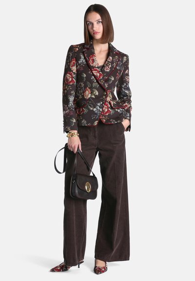 Femme portant un blazer à fleurs et des chaussures assorties, pantalon large foncé, tenant un sac à main noir avec un fermoir doré, debout devant un fond uni.