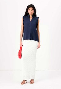 Blusa senza maniche blu navy con spalle arricciate, frontale con bottoni; abbinata a una gonna maxi bianca con texture e porta una borsa rossa.
