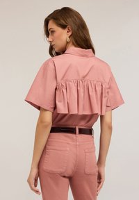 Donna con capelli castani che indossa una camicia rosa a maniche corte con volant e pantaloni abbinati con una cintura scura, in piedi su uno sfondo neutro.