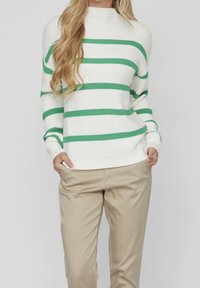 Femme portant un pull blanc à rayures horizontales vertes et un pantalon beige, debout les mains dans les poches devant un fond uni.