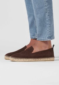 Chaussures slip-on en daim marron avec une semelle en jute tressé, présentant un bout arrondi et des découpes sur les côtés. Portées avec un jean bleu clair retroussé.
