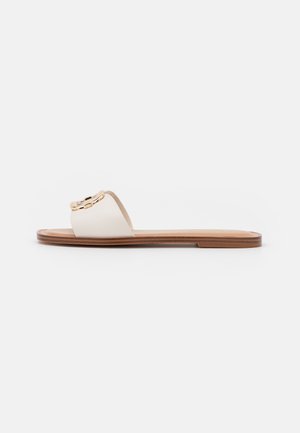 Flad slip sandal med hvid læderrem og guldkædedetalje på en brun sål, vist i profil mod hvid baggrund.