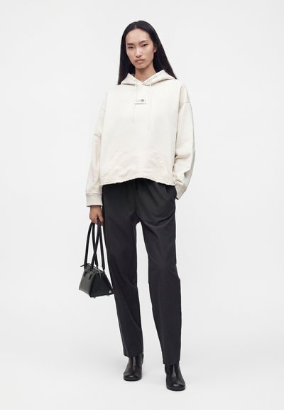 Γυναίκα με μακριά σκούρα μαλλιά φοράει oversized ανοιχτόχρωμο φούτερ με κουκούλα, μαύρο παντελόνι, μαύρα μποτάκια μέχρι τον αστράγαλο και κρατά μια μικρή μαύρη τσάντα στο λευκό φόντο.