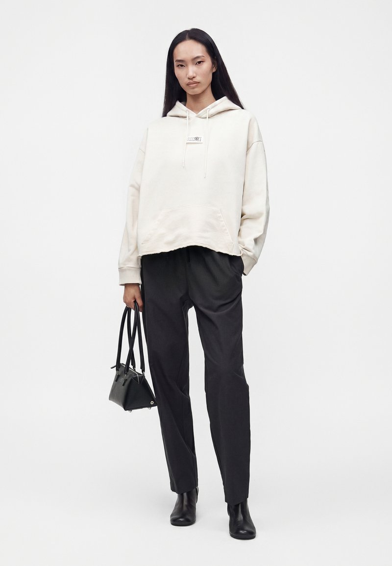 Femme aux longs cheveux foncés porte un sweat à capuche clair et oversize, un pantalon noir, des bottines noires et tient un petit sac à main noir sur fond blanc.
